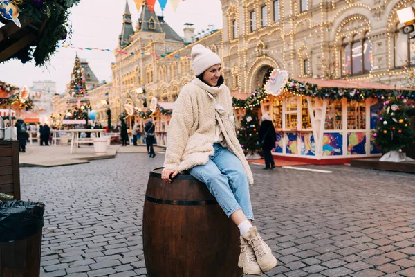 Full body of happy female in warm clothes and hat smiling and looking away while sitting on wooden barrel and looking around at city Christmas fairground