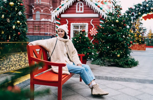 Full body of happy female in warm clothes and hat sitting on red bench and looking away while enjoying Christmas holidays