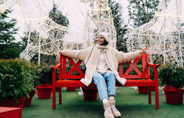 Full length positive young female in warm clothes resting on red cozy bench with arms outstretched against cute horses made of garlands in park decorated for Christmas celebration