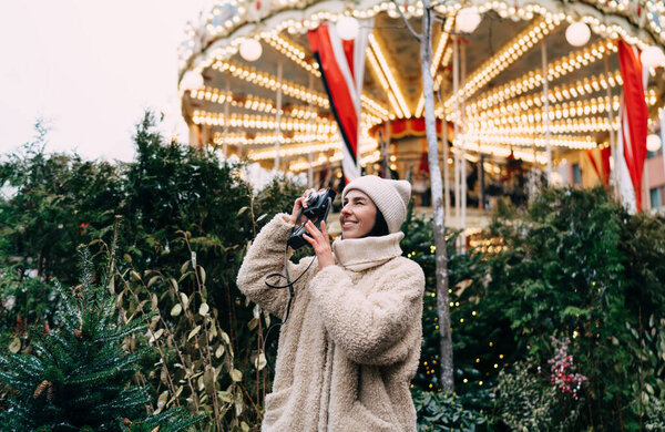 Cheerful woman in warm outwear and hat smiling and taking photo on retro photo camera while walking on decorated Christmas street