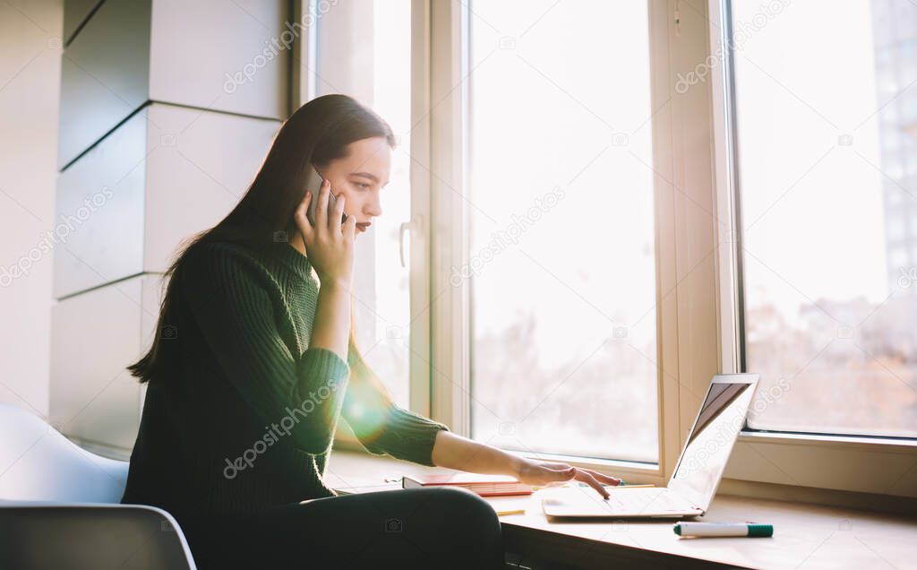 Side view of focused female entrepreneur in casual outfit sitting at windowsill and talking on mobile phone while browsing laptop in cozy workspace