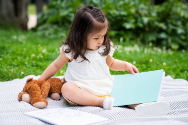 Beyaz elbiseli, siyah saçlı bir çocuk. Pofuduk bir oyuncakla battaniyede oturuyor. Ve parkta yeşil bitkilerle kaplı bir kitap.