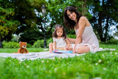 Koyu saçlı, neşeli, mutlu bir kadın beyaz elbiseli, ilginç bir kitabı ve parkta oyuncak ayısı olan bir kızla oturuyor.