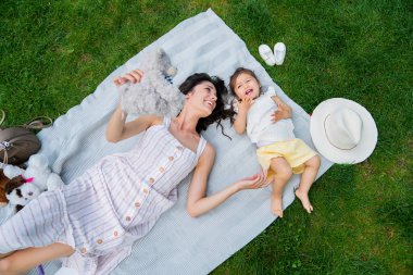 Coşkulu bir annenin sevimli çocuğuyla oynaması ve piknikte battaniyenin üzerinde şapkayla ve ayakkabılarla rahatça yatması.