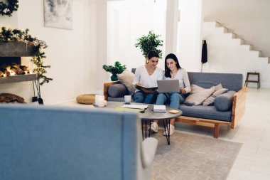 Genç kız arkadaşlar yeni yıl dekorasyonlarıyla dolu odada dizüstü bilgisayarı ve defteriyle rahat koltukta otururken proje yapıyorlar.