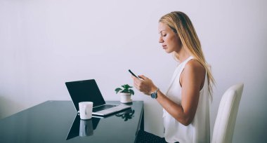 Zarif giyinen, ekrana odaklanan, akıllı telefonlu, netbook ve çağdaş ofisin beyaz duvarına karşı lezzetli içeceklerle dolu bir masada oturan düşünceli, kendine güvenen bir kadının yan görüntüsü.