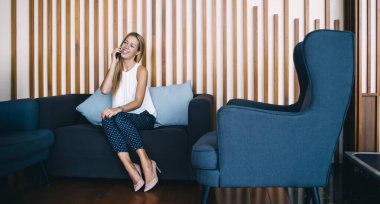 Tam vücut modaya uygun sarışın rahat ışık odasında kanepede oturuyor ve akıllı telefondan mutlu bir şekilde gülümsüyor.