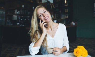 Zarif, kendine güvenen beyaz gömlekli, nefis bir içecekle kameraya gülümseyen bir kadın vazoda parlak sarı çiçeklerle birlikte, çağdaş kahve evinin bulanık iç yüzüne karşı oturuyor.