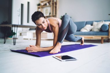 Spor giyimli çıplak ayaklı kadın modern apartmandaki tabletle online yoga dersine katılırken minder üzerinde yoga egzersizi yapıyor.