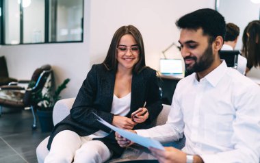 Gözlüklü, akıllı telefonlu, gülümseyen bir iş kadını resmi giysiler içinde erkek iş arkadaşıyla koltukta oturuyor ve çağdaş çalışma alanında tartışıyor.