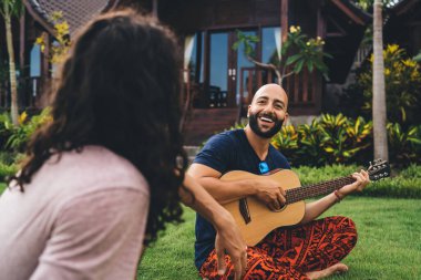 Mutlu, sakallı, sıradan giyinmiş yeşil çimenlerde oturan ve arkadaşlarıyla bahçede gitar çalan etnik bir adam.