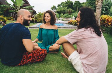 Parktaki yeşil çimlerde oturan ve birbirleriyle konuşurken eğlenen mutlu yetişkin arkadaş grubu.