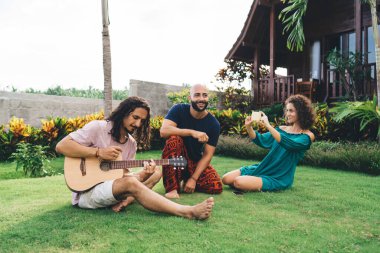 Tam vücut iyimser çok ırklı arkadaşlar, günlük kıyafetleriyle yeşil çimlerin üzerinde oturuyorlar ve hafta sonu birlikte gitar çalıyorlar.