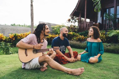 Serbest giyinen çok ırklı arkadaşlardan oluşan tam bir grup yeşil çimenlerde oturup birlikte gitar çalıyor.