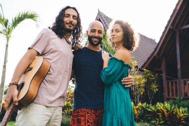 Düşük açılı gülümseyen çok ırklı arkadaş grubu gitarla takılırken kameraya bakıyor ve sarılıyorlar.