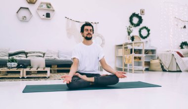 Sakin sakallı, gözleri kapalı bir erkek spor minderinin üzerinde oturuyor ve Padmasana 'da meditasyon yapıyor. Evdeki aydınlık oturma odasında.