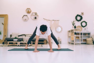 Spor kıyafetleri içinde çıplak ayaklı bir erkek mindere eğilip Prasarita Padottanasana yaparken öne doğru eğiliyor.