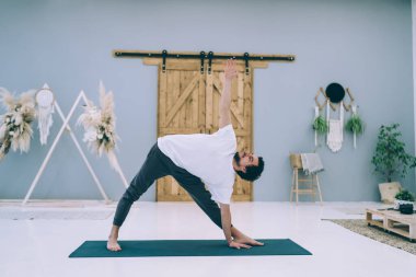 Spor kıyafetleri içinde sakallı bir erkek mindere çıkıp Trikonasana çalışırken modern tarzda rahat bir odada ısınıyor.