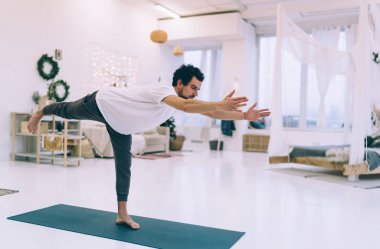 Spor kıyafetleri içinde sakallı bir erkek, rahat oturma odasında yoga yaparken Warrior III pozu veriyor.