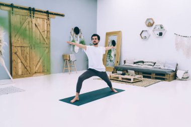 Çıplak ayaklı sakallı bir erkek, Crescent Lunge Twist egzersizi yapıyor. Dairedeki geniş odada ellerini spor paspasına uzatmış.