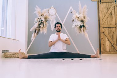 Karizmatik sakallı, siyah saçlı spor elbiseli, evde yoga seansı sırasında çapraz bacak egzersizi yapan bir erkek.