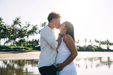 Aşk dolu hamile bir çiftin, güneş ışığı altında ıslak kumlu sahilde romantik zaman geçirirken tutkuyla öpüştüğünün yan görüntüsü.