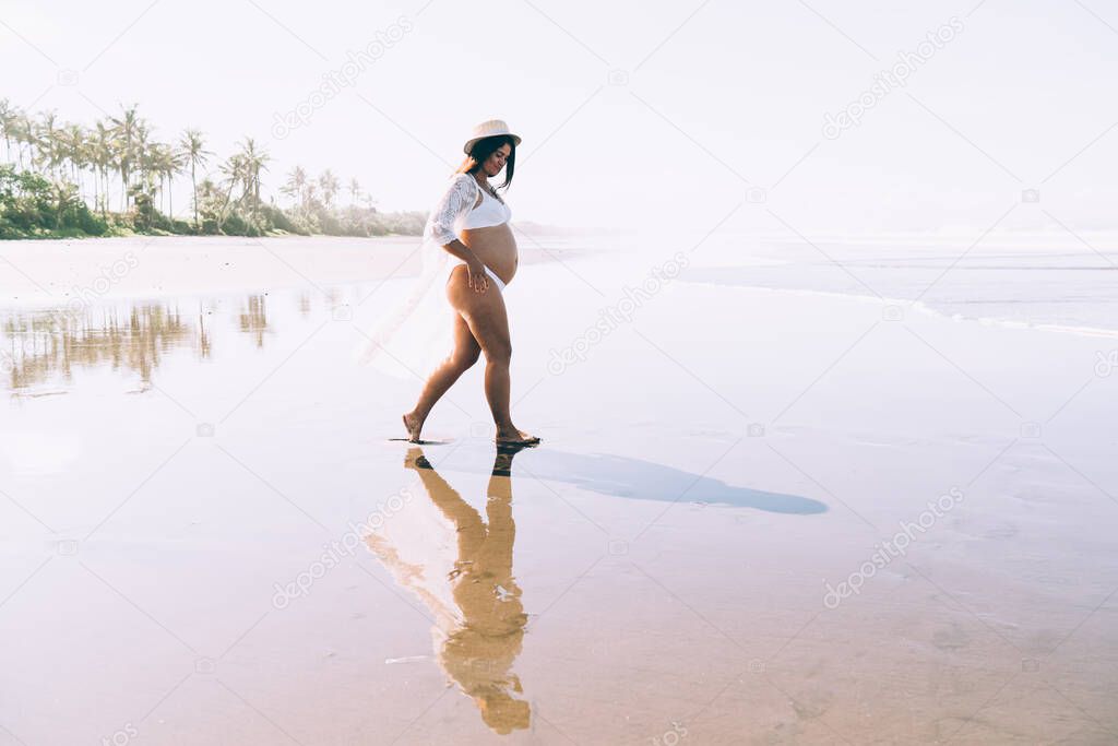 Side view of positive young pregnant female in stylish swimwear and hat walking on wet sandy beach towards waving ocean during summer holidays in tropical country