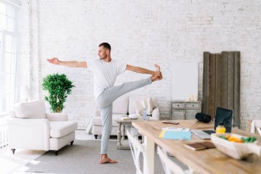 Spor kıyafetleri içinde çıplak ayaklı bir adam modern oturma odasında Ayakta Ayakta Ayakta Durma pozu çalışırken gözlerini kaçırıyor.