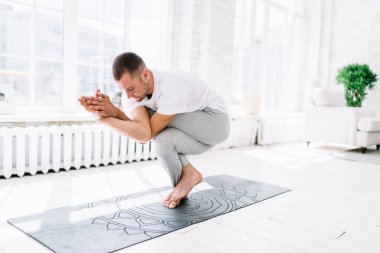Çıplak ayaklı erkek, spor kıyafetleri içinde Eagle Crunch pozisyonunda yoga ve bacak üzerinde denge kurarken minder üzerinde duruyor.