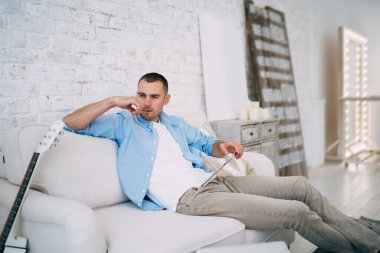 Düşünceli sakallı bir adam günlük kıyafetler giyiyor ve tablet okurken gözlük tutuyor ve uzaktan çalışma sırasında rahat bir koltukta oturuyor.