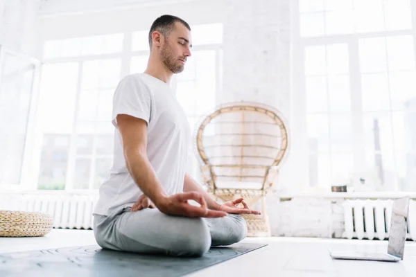 stock image Calm caucasian man with closed eyes sitting in lotus pose meditating feeling mindfulness and spirituality, handoms hipster guy using laptop computer for training yoga online staying on isolation