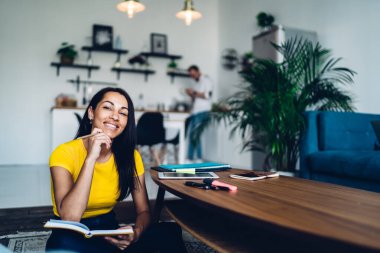 Laptop ve renkli kalemlerle günlük kıyafetli genç etnik kız öğrenci kahve masasının yanında oturmuş üniversite dersleri için notlar hazırlarken kameraya bakıyor.