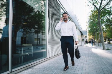 Elinde modaya uygun evrak çantasıyla orta yaşlı bir işadamı telefon kullanarak şehir caddesinde yürüyor ve iş görüşmeleri yapıyor. Cep telefonuyla cep telefonuyla geziyor. Cep telefonu konuşmaları için gezen kendine güvenen bir girişimci.