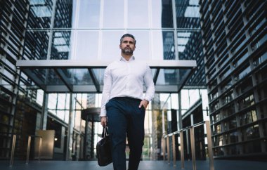 Tasarımlı orta yaşlı bir işadamı şık bir evrak çantasıyla şirketinin yanında yürüyen beyaz erkek bankacı finans bölgesinde bulunan ofis binasından dışarı çıkıyor.