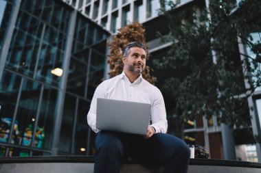 Düşünceli, olgun bir işadamı, modern dizüstü bilgisayarında uzaktan yatırım yaparken, düşünceli, düşünceli, 40 yaşında beyaz bir bankacı elinde ağlarla pazarlama üzerine kafa yoruyor.