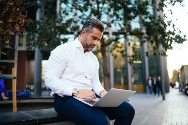 Olgun erkek tüccar online iş mesajları yapıyor şehir ortamında dışarıda oturuyor, orta yaşlı girişimci modern dizüstü bilgisayara web dosyaları yüklemek için 4G kablosuz internet bağlantısı kullanıyor