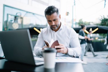 Profesyonel Kafkasyalı yönetici, uzaktaki bir kafede kişisel planlamacı kullanıyor. Modern laptop teknolojisi ile masada oturuyor ve bilgi notlarını analiz ediyor. Beyaz gömlekli iş adamı çalışıyor.
