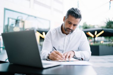 Olgun girişimci ders kitabına notlar yazarak modern netbook ile kaldırım kafeteryasında oturuyor ve orta yaşlı gururlu CEO 'ların finans bölgesinde iş planlaması yapıyor.