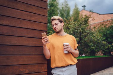 Düşük açılı, düşünceli, turuncu gömlekli, sıcak içecekli sarışın bir erkek. Ekrana odaklanmak ve akıllı telefon kullanmak için. Sokakta dikilip park alanındaki modern binanın ahşap çizgili duvarına yaslanırken.