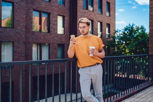 都会の金属製の柵に寄りかかってコーヒーを飲みながら携帯電話を真剣に街づくりに反対しているスタイリッシュな濃縮若い男性 — ストック写真