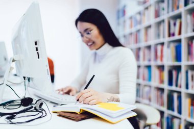 Koyu renk saçlı, gözlüklü, beyaz bilgisayarlı ve defterine yazı yazan, kütüphanedeki masada oturan mutlu kadının yumuşak odağı.