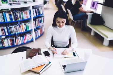 Resmi giysiler giymiş, elinde kalem tutan ve elinde kitaplarla masa başında otururken not defterine odaklanan düşünceli etnik yetişkin bir kadından. Gündüz vakti bulanık arka planda hafif modern kütüphanede içecekleri alıp götürüyor.