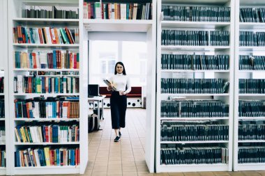 Beyaz balıkçı yaka kazaklı, siyah pantolonlu, elinde klasörler ve kağıt bardakla üniversite kütüphanesinde kitaplıklarla kapıdan içeri giren Asyalı bir kadın.