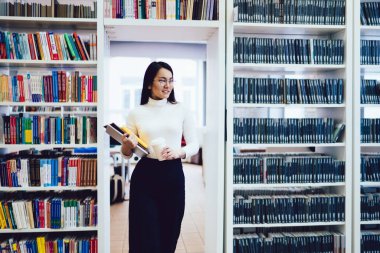 Aktif stil sahibi siyah pantolonlu Asyalı kadın ve beyaz balıkçı yakalı, elinde defterler ve açık kütüphanedeki kitaplıklar arasında yürüyen tek kullanımlık beyaz kahve fincanı var.