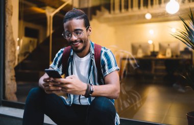 Gülümseyen Afro-Amerikan hipster adam akıllı telefon uygulaması ile kontrol ediyor boş zamanlarında sokakta oturuyor, pozitif koyu tenli erkek 4G bağlantısından memnun ve medya içeriğini paylaşıyor.