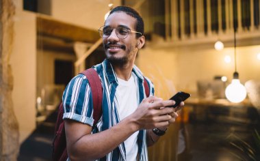 Mutlu Afro-Amerikan adam cep telefonu tutuyor, mesajlar gönderiyor ve başka tarafa bakıyor, pozitif yakışıklı, koyu tenli hipster adam akıllı telefon ve 4G bağlantısı ile iletişim kuruyor.