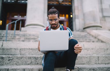 Düşük açılı sakallı, gözlüklü etnik adam internette internette gezinirken gün ışığında merdivenlerde oturuyor.
