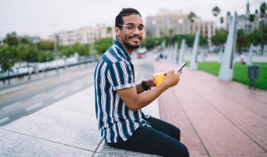 Moda kıyafetli, gözlüklü, tek kullanımlık kahve bardaklı, akıllı telefondan mesajlaşan, şehrin caddesinde yol kenarında oturan sakallı etnik adamın yan görüntüsü.