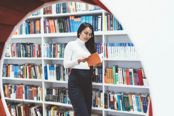 专心致志的亚洲女学生 身穿休闲装 戴着眼镜 站在高校图书馆书架上看书 独自做作业 — 图库照片