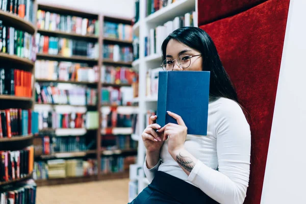 긍정적 아시아 여성이 책으로 가리고 도서관에서 카메라를 창조적 의자에서 — 스톡 사진